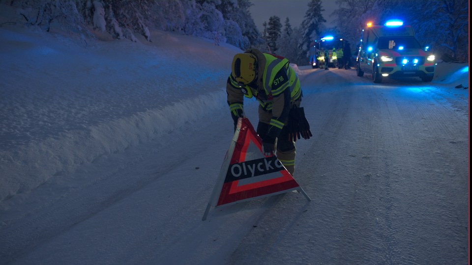 Räddningstjänsten placerar ut en varningsskylt med texten olycka på en snöig väg. I bakgrunden syns blåljusfordon.