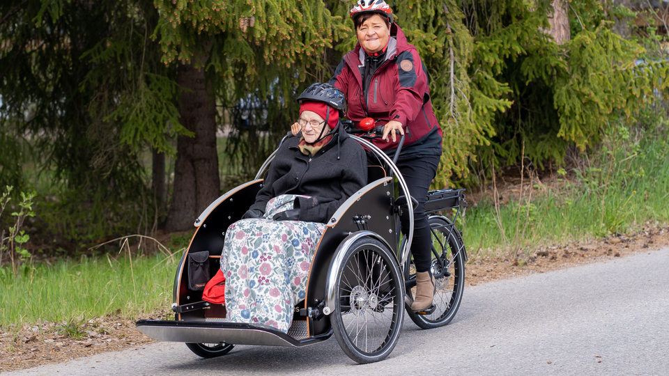 Kvinna som jobbar inom äldreomsorgen skjutsar äldre kvinna på en parcykel.