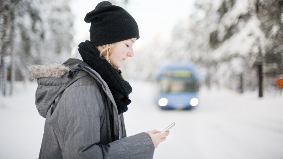 En tjej tittar på sin telefon i ett snöigt landskap och en buss kör i bakgrunden.